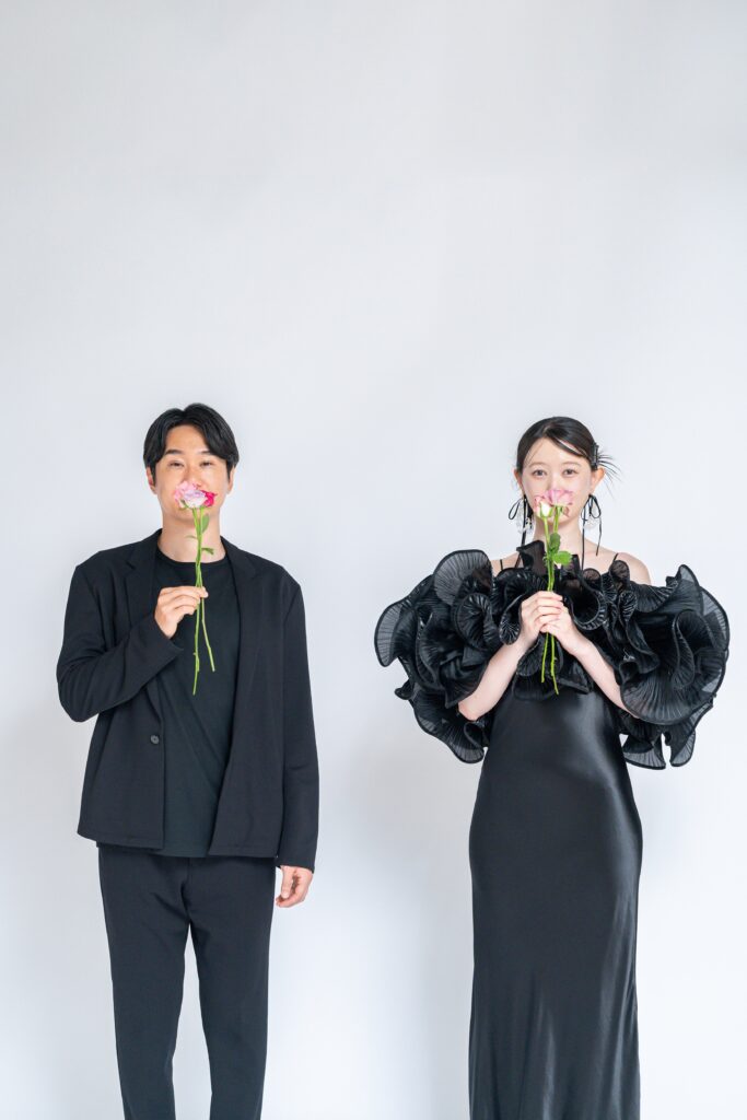 黒のジャケットスタイルとフリルドレスで、ピンクのバラを持つおふたり