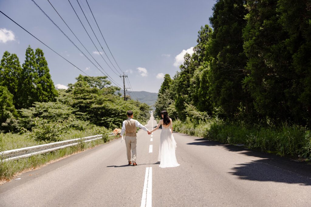 自然の一本道を手をつないで歩く新郎新婦のロケーションフォト
